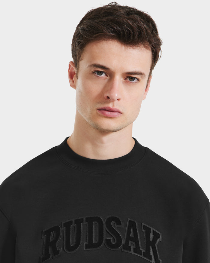 A young man with short brown hair wears the Rudsak MILOS - 6525646 Black men’s crewneck sweatshirt with bold RUDSAK lettering on the chest, looking directly at the camera with a neutral expression.