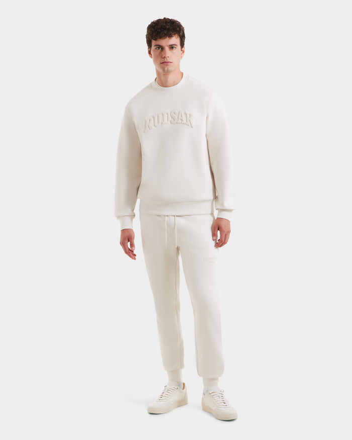 A man stands facing forward in Rudsak’s MILOS - 6525646 Vanilla men’s crewneck sweatshirt and matching jogger pants, both in off-white premium cotton blend with embossed branding, styled minimally against a plain white background.