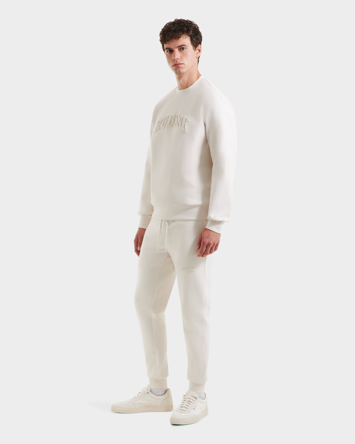 A man wears the Rudsak MILOS - 6525646 Vanilla set—an all-white, minimalist crewneck sweatshirt with embossed text and matching jogger pants—styled with white sneakers against a plain white background.