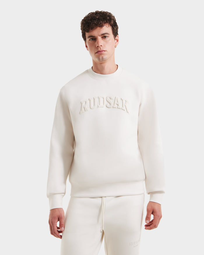 A young man in the MILOS - 6525646 Vanilla men’s premium Rudsak sweatshirt and matching sweatpants set with embossed Rudsak text stands against a plain white background.