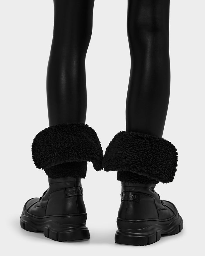 A person wearing shiny black leggings and Rudsak MODENA - 8225024 Black chunky winter boots with large, folded-down, faux shearling cuffs, shown from behind against a plain background.