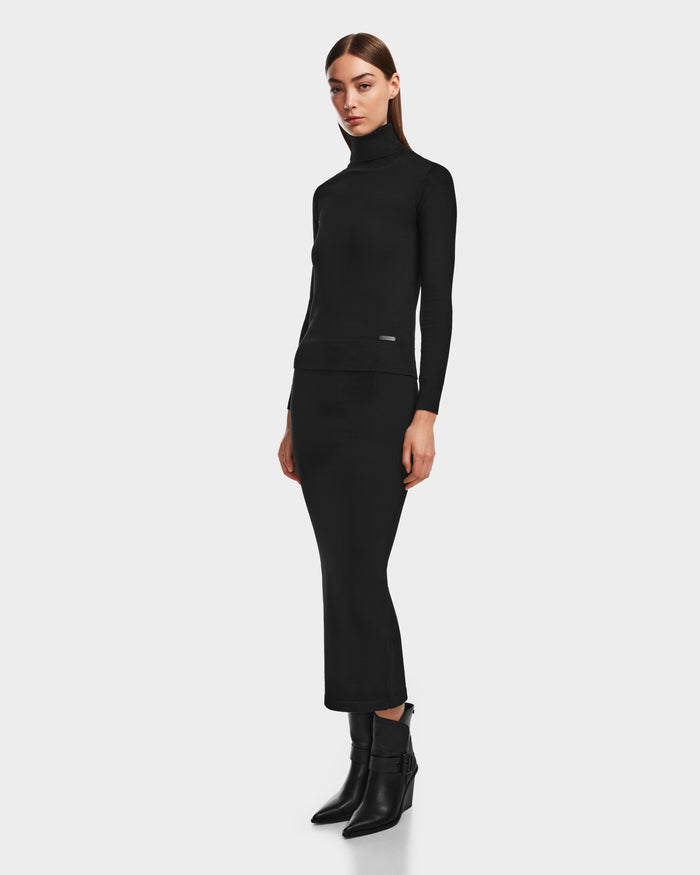 A woman with straight brown hair and a neutral expression stands against a white background, wearing the Rudsak MONICA - 8524516 Black fitted turtleneck and knit skirt, paired with black heeled ankle boots.