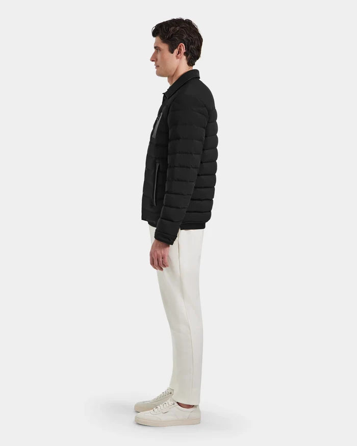 A man stands in profile against a plain background, wearing the Rudsak NICHOLAS - 6125040 Black quilted shirt jacket—an essential for any men’s wardrobe—paired with white pants and sneakers.