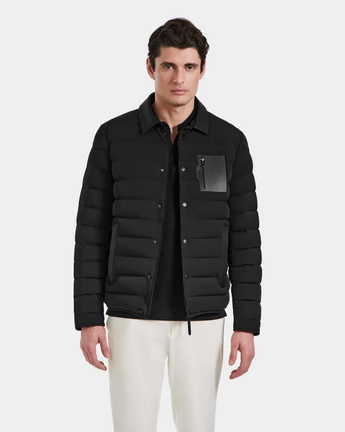A man faces forward in the Rudsak NICHOLAS - 6125040 Black quilted shirt jacket, made with RDS-certified down and a zippered chest pocket, paired with white pants against a plain light background—a modern outerwear look.