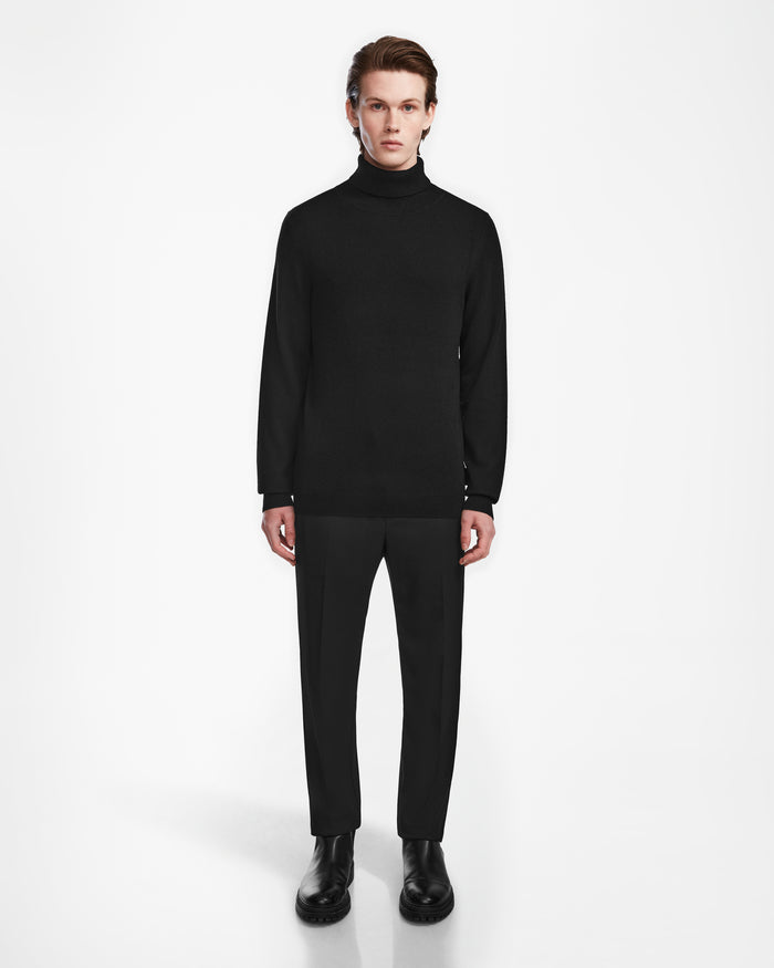A person with light skin and brown hair stands facing forward against a white background, wearing the Rudsak NICOLAS - 6524528 Black turtleneck sweater, black dress pants, and black shoes.