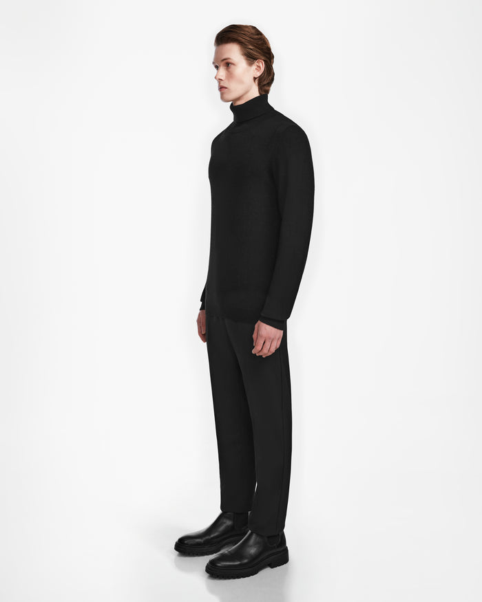 A person with light skin and medium-length brown hair stands in profile, wearing Rudsak’s NICOLAS - 6524528 Black luxury turtleneck sweater with black pants and shoes, set against a plain white background.