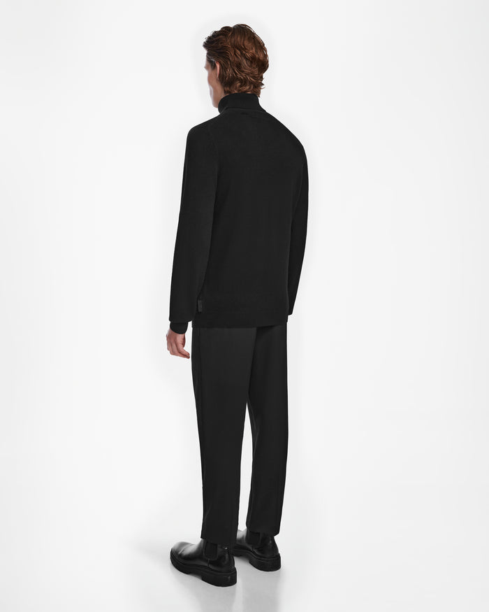 A person with short brown hair faces away, wearing Rudsak’s NICOLAS - 6524528 Black men’s cashmere turtleneck sweater and black pants, creating a sleek look against a plain white background.