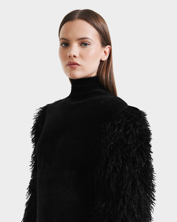 A woman with straight brown hair wears the Rudsak NOEL - 8525571 Black knit sweater with faux fur sleeves, standing against a plain light background and looking slightly to the side.