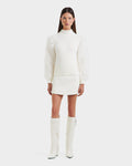 A woman faces forward in winter style, wearing the Rudsak NOEL - 8525571 Vanilla mock-neck sweater with faux fur sleeves, a white mini skirt, and tall white heeled boots. She has straight brown hair and stands against a plain white background.
