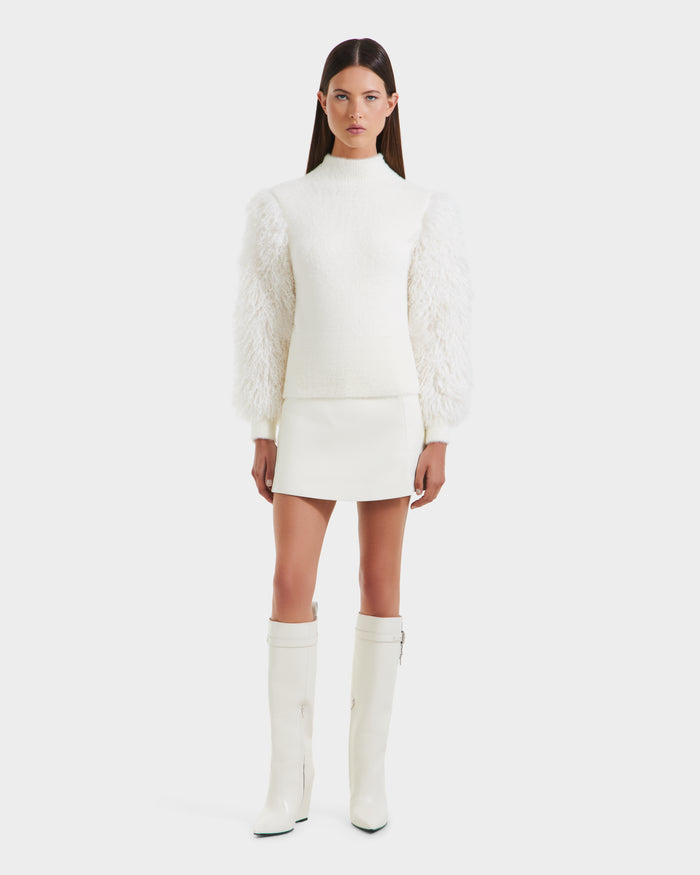 A woman faces forward in winter style, wearing the Rudsak NOEL - 8525571 Vanilla mock-neck sweater with faux fur sleeves, a white mini skirt, and tall white heeled boots. She has straight brown hair and stands against a plain white background.