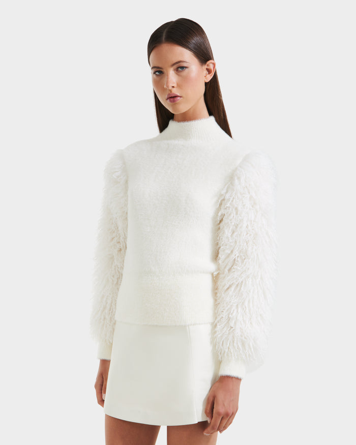 A woman with straight brown hair and a neutral expression models the Rudsak NOEL - 8525571 Vanilla women’s mock-neck sweater with faux fur sleeves, paired with a matching white skirt, against a plain light background.