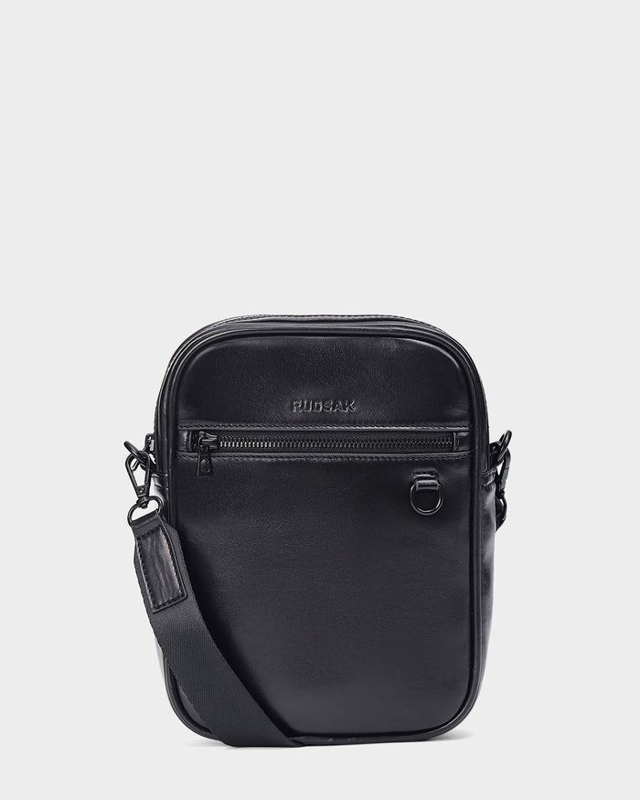 The Rudsak NORMAN L - 9323503 Black is a unisex crossbody bag with a front zip pocket, embossed logo, metal ring detail, and adjustable strap, shown against a plain white background.