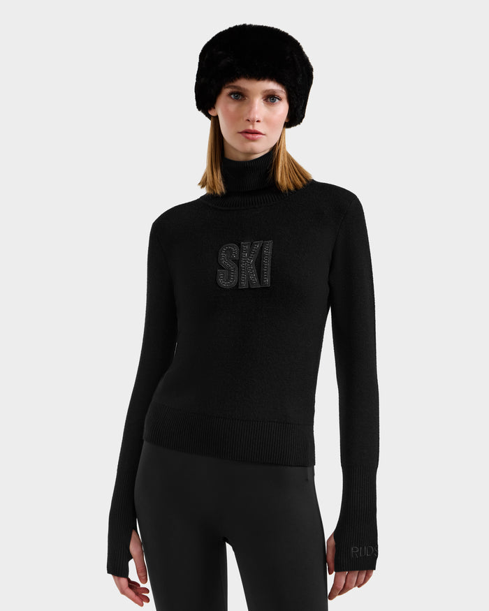 A woman models the Rudsak NOVIE - 8525659 Black ski sweater with "SKI" on the front, paired with black fitted pants and a furry hat, creating an effortlessly chic après-ski look against a light gray background.