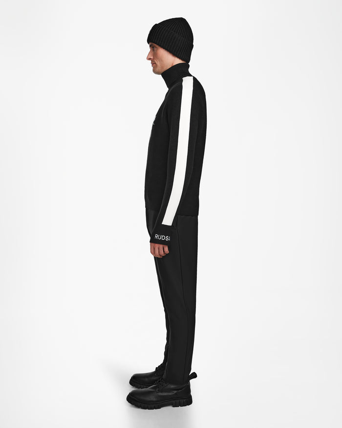 A person wears the Rudsak OLIVIER - 6524545 Black après-ski-inspired sweater with a white stripe on the sleeve, styled with black beanie, pants, and boots, standing in profile against a plain white background.
