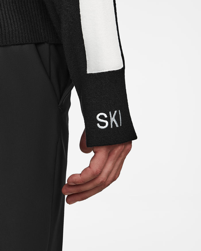 A close-up of a person’s arm in the OLIVIER - 6524545 Black men’s ski turtleneck sweater by Rudsak, featuring “SKI” embroidered on the sleeve and paired with black pants—a stylish choice for any winter wardrobe or après-ski occasion.