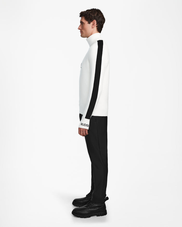 A man stands in profile wearing the Rudsak OLIVIER - 6524545 Optic White ski turtleneck with black striped sleeves, paired with black pants and boots, creating a sleek winter look against a plain white background.