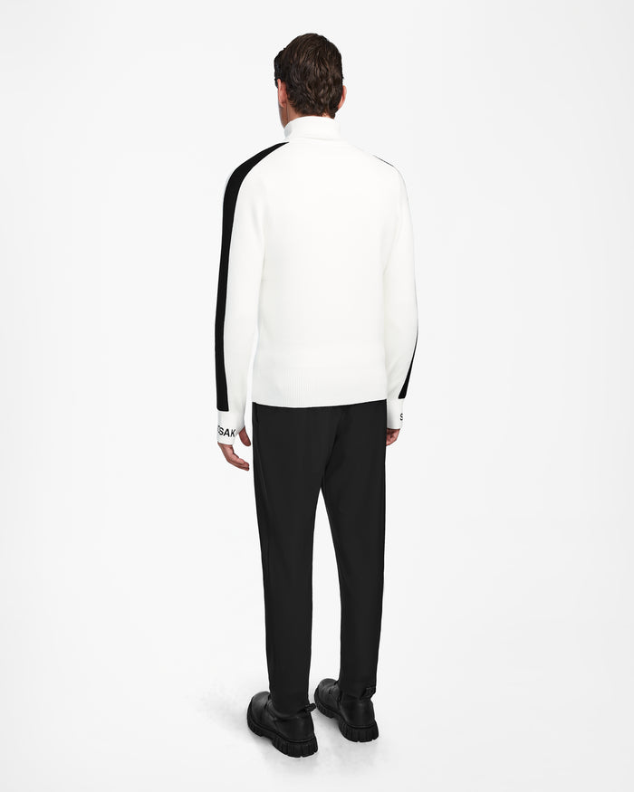 A man stands facing away in the Rudsak OLIVIER - 6524545 Optic White mens ski turtleneck with black stripes, paired with black pants and shoes—ideal winter wear—set against a plain white background.