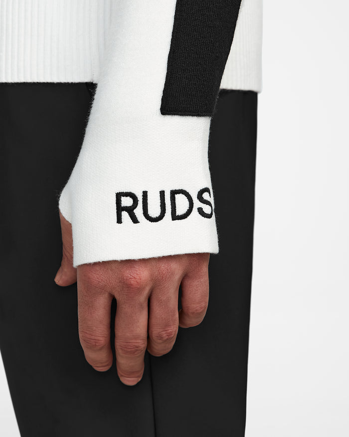 A close-up of a person’s left hand wearing the OLIVIER - 6524545 Optic White men’s ski turtleneck by Rudsak, featuring black RUDS text on the cuff, a black section on the sleeve, and paired with black pants—ideal for winter.