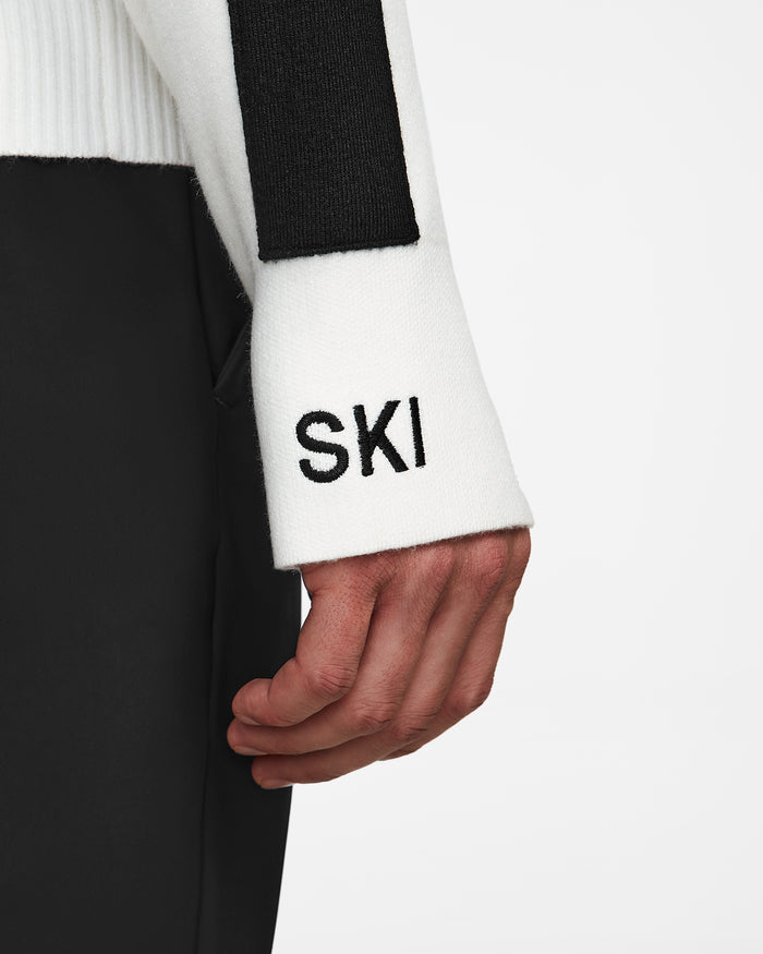 A close-up of a hand resting by the side, wearing the Rudsak OLIVIER - 6524545 Optic White sweater with SKI embroidered in black, paired with black pants against a plain white background.