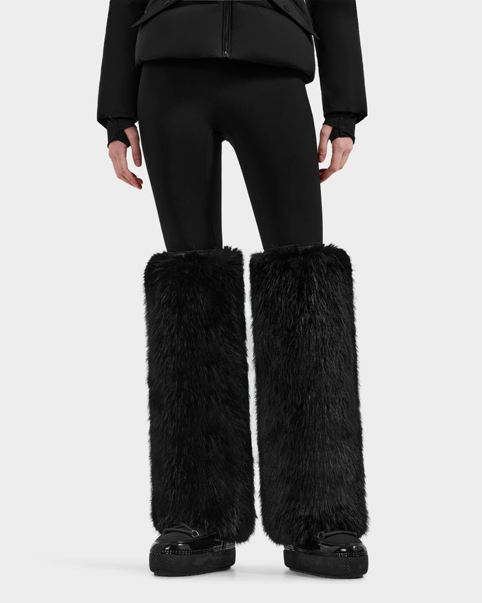 Clad in Rudsak’s PAIGE - 8425534 Black leggings and jacket with oversized faux fur leg warmers, this striking look stands out against a light background—perfect for après-ski style.