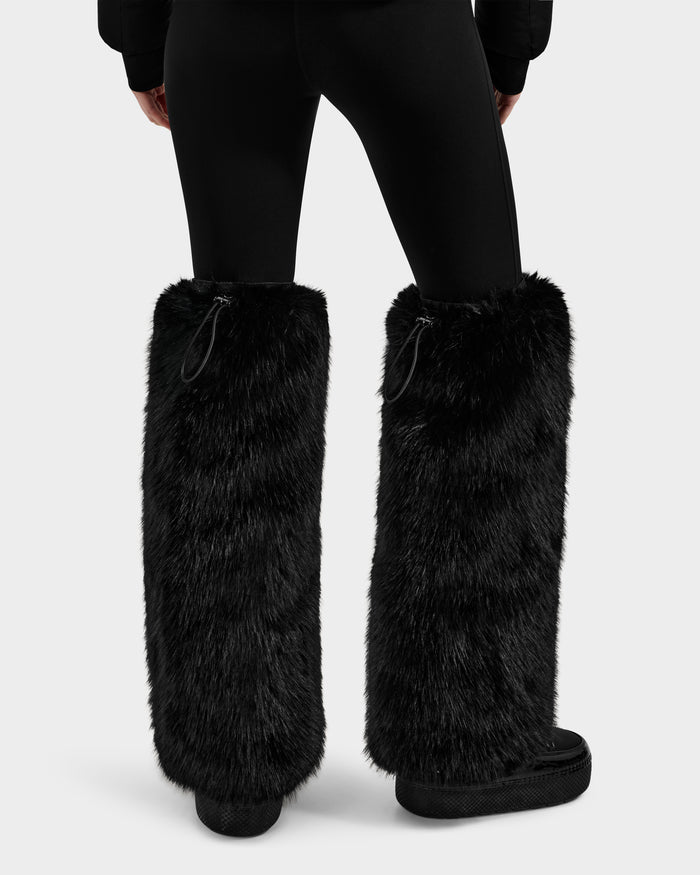 A person displays après-ski chic in Rudsak's PAIGE - 8425534 Black faux fur leg warmers over black leggings and platform shoes, photographed from the back against a plain light background.
