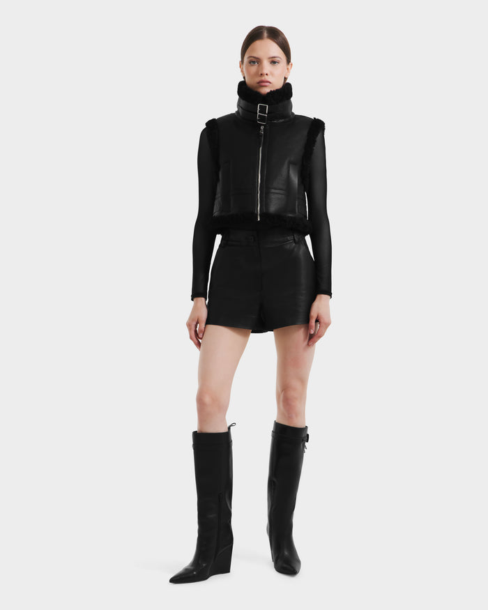 A woman stands facing forward against a plain background, wearing the Rudsak PAISLEY - L125516 Black cropped vegan leather vest, paired with a black long-sleeve top, high-waisted shorts, and knee-high black wedge boots.