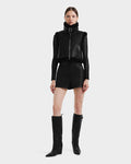 A woman stands facing forward against a plain background, wearing the Rudsak PAISLEY - L125516 Black cropped vegan leather vest, paired with a black long-sleeve top, high-waisted shorts, and knee-high black wedge boots.