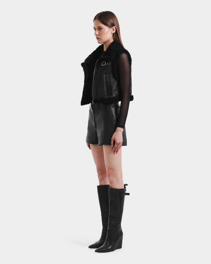 A woman in profile wears the Rudsak PAISLEY - L125516 Black faux shearling vest, a black leather mini skirt, sheer long-sleeve top, and black wedge knee-high boots, standing against a plain white background.