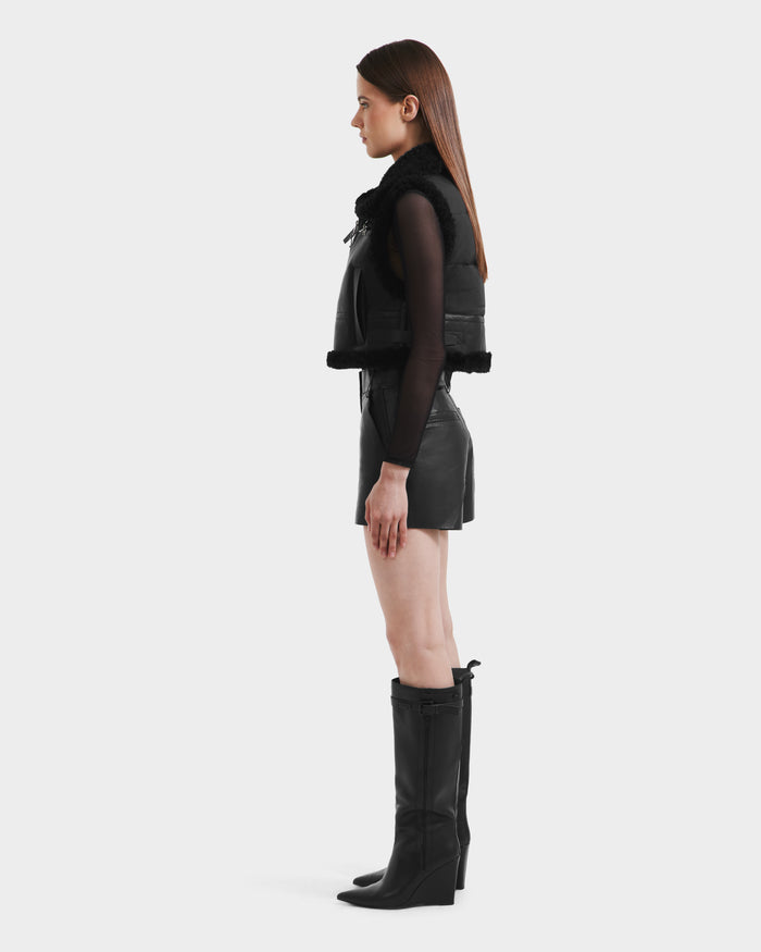 A woman stands in profile against a plain background, wearing the Rudsak PAISLEY - L125516 Black cropped vegan leather vest with fur trim, a black long-sleeve top, black leather shorts, and knee-high black boots.