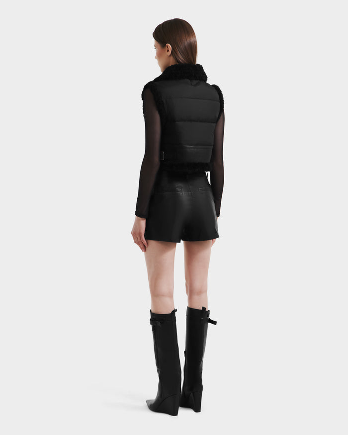 A woman faces away wearing Rudsak's PAISLEY - L125516 Black vegan leather cropped vest, paired with high-waisted black shorts, a sheer long-sleeve top, and knee-high heeled boots against a plain light background.