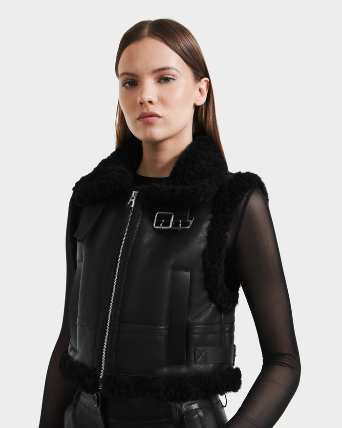 A woman wears the Rudsak PAISLEY - L125516 Black women's cropped vegan leather vest with faux shearling collar and zipper over a sheer black long-sleeve top, standing against a plain light background.
