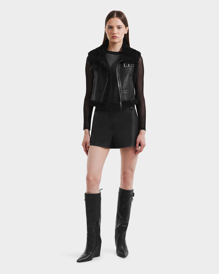 A woman stands facing forward, wearing the Rudsak PAISLEY - L125516 Black vegan leather vest with faux shearling collar, over a sheer black top, black shorts, and knee-high pointed-toe block-heel boots.