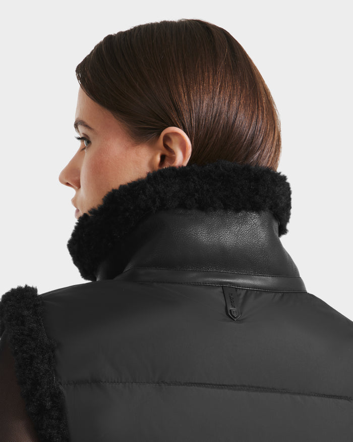 A person with straight brown hair is shown from behind and in profile, wearing the Rudsak PAISLEY - L125516 Black women’s cropped vegan leather vest with a high textured collar and leather trim, against a plain light background.
