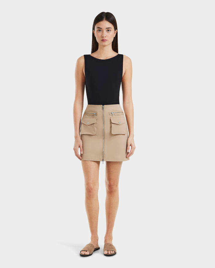 A woman stands facing forward wearing Rudsaks PAMINA - 8525025 Almond vegan leather mini skirt with utility pockets and zippers, a sleeveless black top, and tan slide sandals against a plain white background.