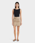 A woman stands facing forward wearing Rudsaks PAMINA - 8525025 Almond vegan leather mini skirt with utility pockets and zippers, a sleeveless black top, and tan slide sandals against a plain white background.