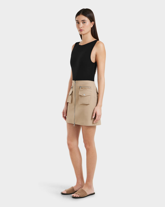 A woman with straight dark hair and a neutral expression wears the Rudsak PAMINA - 8525025 Almond beige vegan leather mini skirt with utility pockets and zipper details, paired with a sleeveless black top and black slide sandals against a plain background.