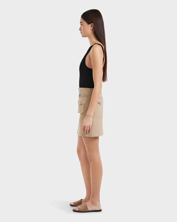 A woman stands in profile against a plain white background, wearing a sleeveless black top, a women’s mini skirt with utility pockets, and Rudsak PAMINA - 8525025 Almond brown flat sandals.
