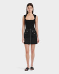 A woman faces forward in the Rudsak PAMINA - 8525025 Black mini dress with a square neckline, front zipper, and utility pockets, paired with black slide sandals against a plain white background.