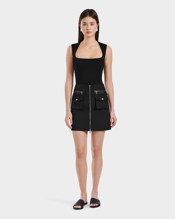 A woman faces forward in the Rudsak PAMINA - 8525025 Black mini dress with a square neckline, front zipper, and utility pockets, paired with black slide sandals against a plain white background.