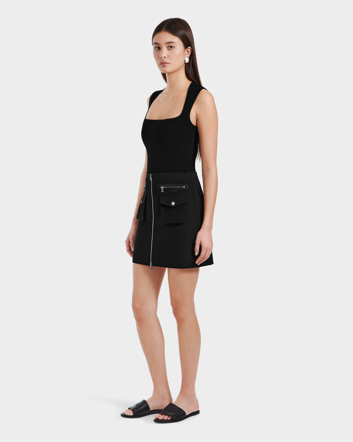 A woman with long brown hair wears the Rudsak PAMINA - 8525025 Black sleeveless dress with utility pockets and a zipper, paired with black slide sandals. She stands slightly turned against a plain white background.