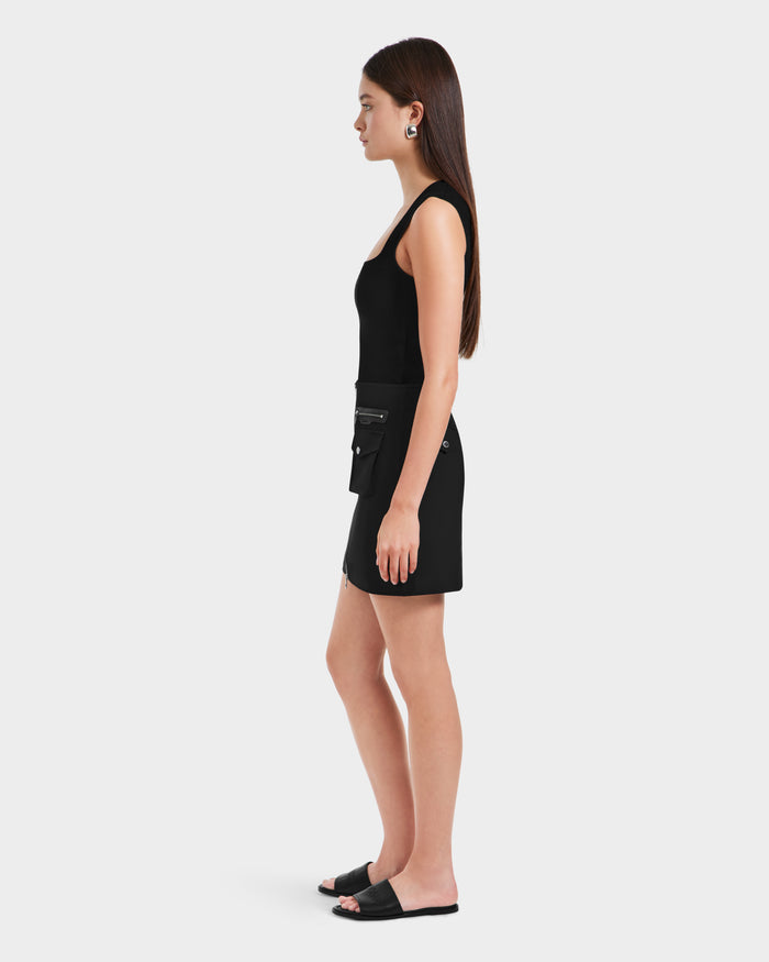 A woman stands in profile against a plain background, wearing the Rudsak PAMINA - 8525025 Black women’s vegan leather mini skirt with utility pockets and zippers, a black sleeveless top, black slide sandals, hoop earrings, and her straight hair worn down.