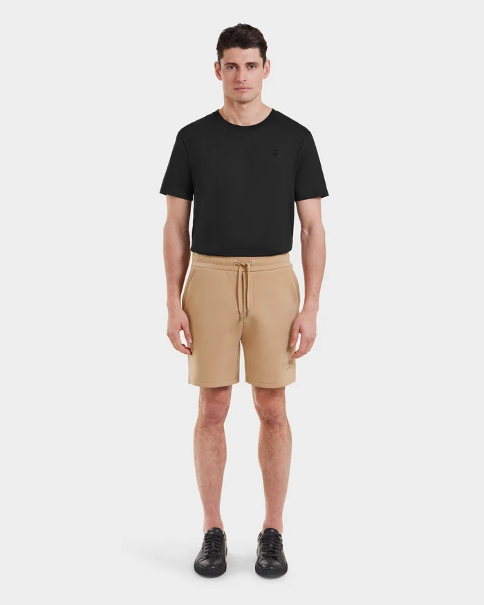 A man stands facing forward in a Rudsak PASCAL - 6524022 Almond t-shirt, casual drawstring shorts, and black sneakers against a plain white background.