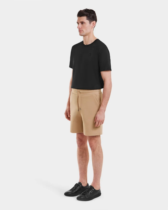 A man stands against a plain white background, wearing Rudsak PASCAL - 6524022 Almond shorts, a black T-shirt, and black sneakers. He looks forward with his arms relaxed by his sides.