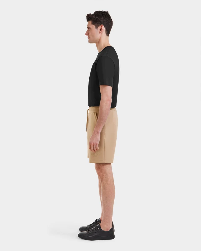 A man stands in profile against a plain background, wearing a black t-shirt, Rudsak shorts, black sneakers, and the Pascal - 6524022 Almond shoes by Rudsak.