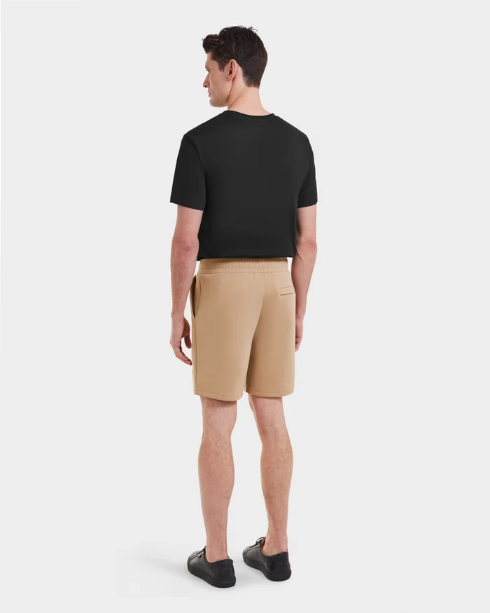 A man faces away, wearing Rudsak's PASCAL - 6524022 Almond beige men's casual shorts, a black short-sleeve t-shirt, and black shoes against a plain white background.