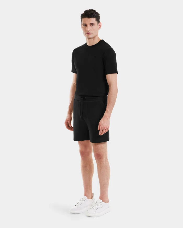 A man faces forward against a light gray background, wearing a black short-sleeve t-shirt, Rudsak PASCAL - 6524022 Black mens double jersey shorts with a stretch comfort waistband, and white sneakers.