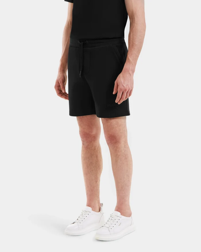 A person stands against a plain background wearing a black t-shirt, Rudsak PASCAL - 6524022 Black mens jersey shorts with a stretch waistband, and white sneakers. The image captures the figure from the shoulders down.