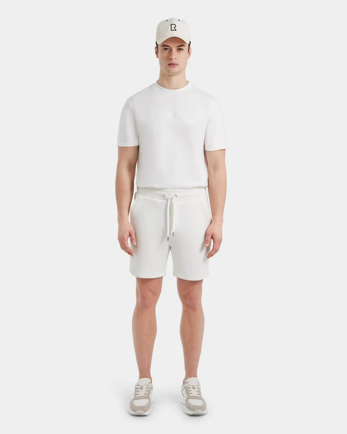 A person stands facing forward in the Rudsak PASCAL - 6524022 ICELAND men’s jersey shorts with a stretch waistband, paired with a white cap, white short-sleeve shirt, and beige and white sneakers against a plain white background.