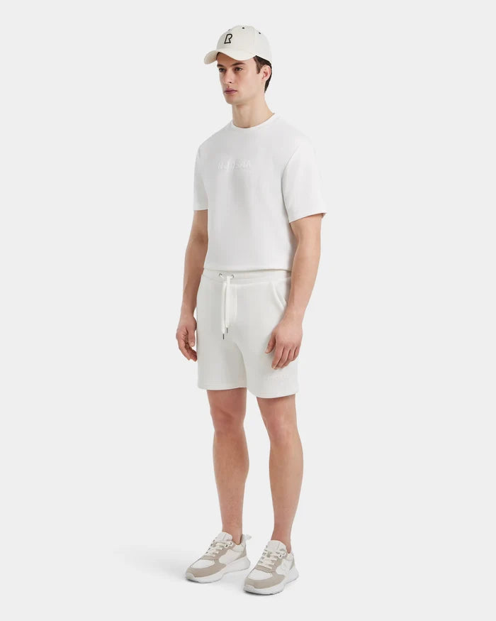 A man stands against a light background wearing a white cap, white t-shirt, Rudsak PASCAL - 6524022 ICELAND shorts with a stretch comfort waistband, and white sneakers with beige accents.