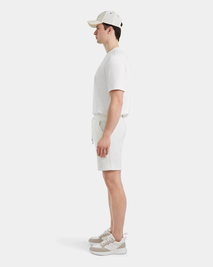 A man stands in profile wearing a white cap, white T-shirt, Rudsak PASCAL - 6524022 ICELAND shorts with a stretch comfort waistband, and beige-and-white sneakers against a plain white background.